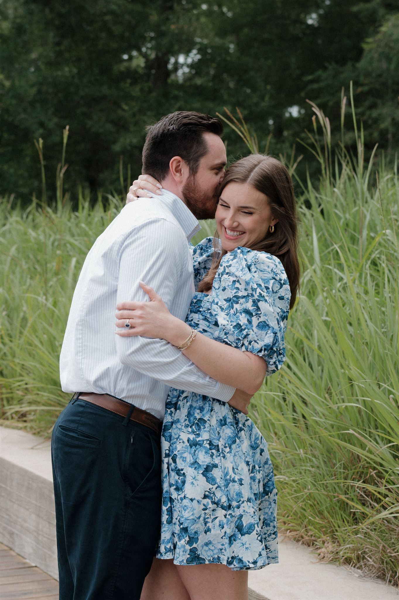 A couple embraces while standing on a wooden boardwalk surrounded by tall grass on a summer day.