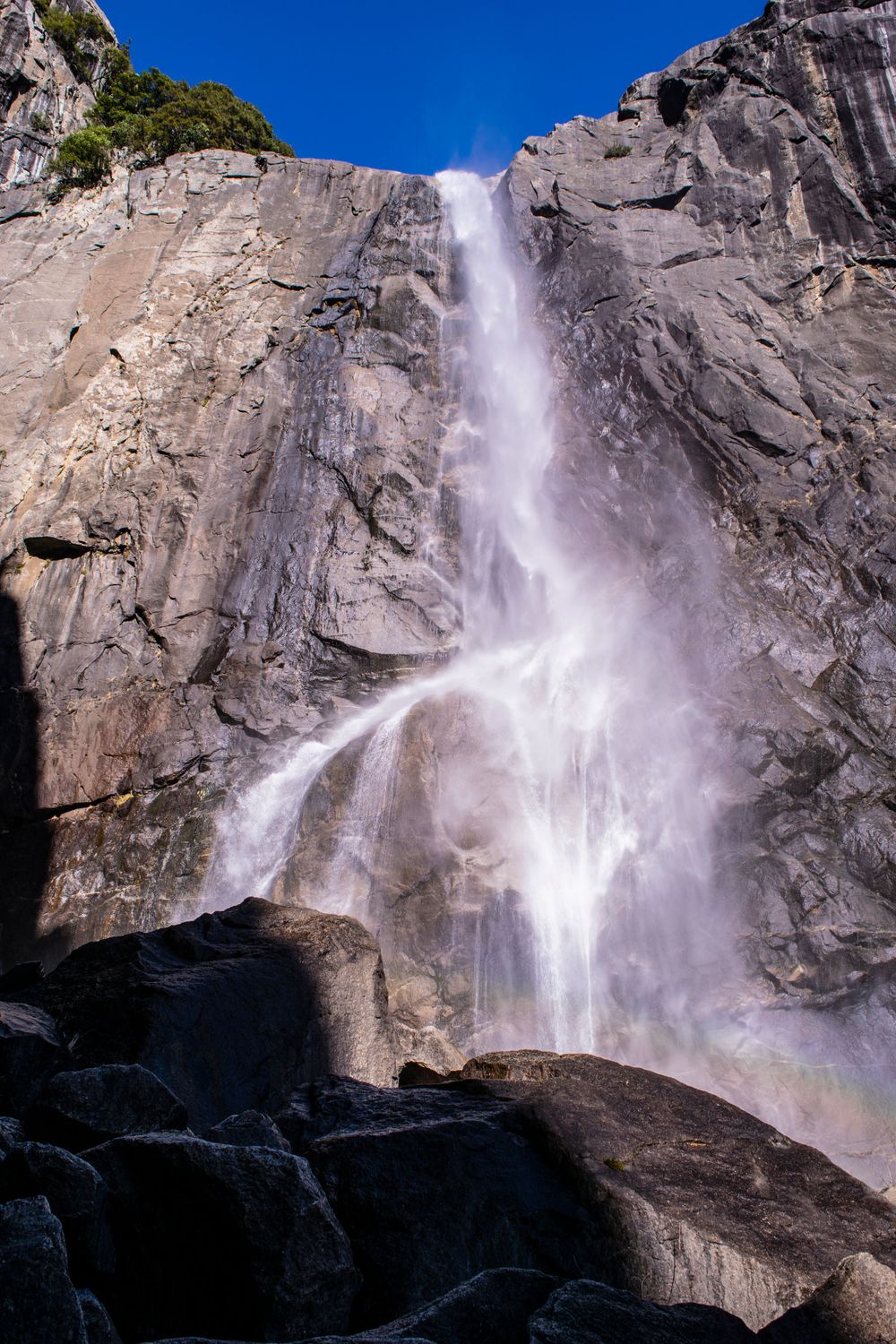 Misty waterfall creates rainbow as it crashes down granite cliff face against bright blue sky.