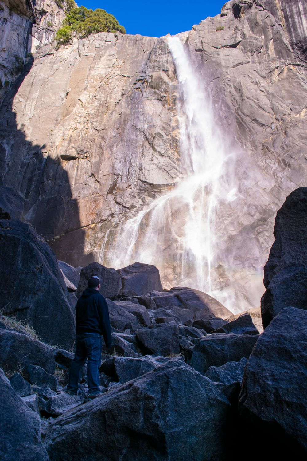 Silhouette stands on rocky terrain viewing powerful waterfall cascading down cliff face.