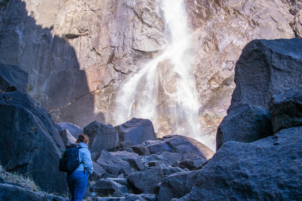 Figure in blue stands among boulders beneath towering waterfall in mountain landscape.