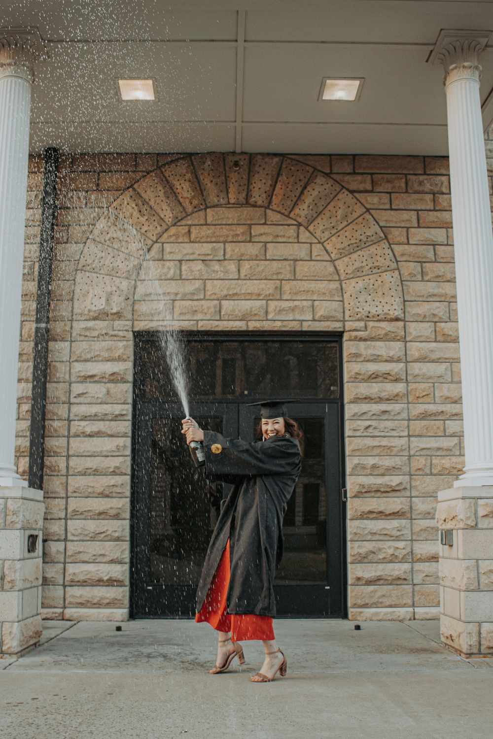 A graduate in academic regalia celebrates under falling water in front of a stone archway.