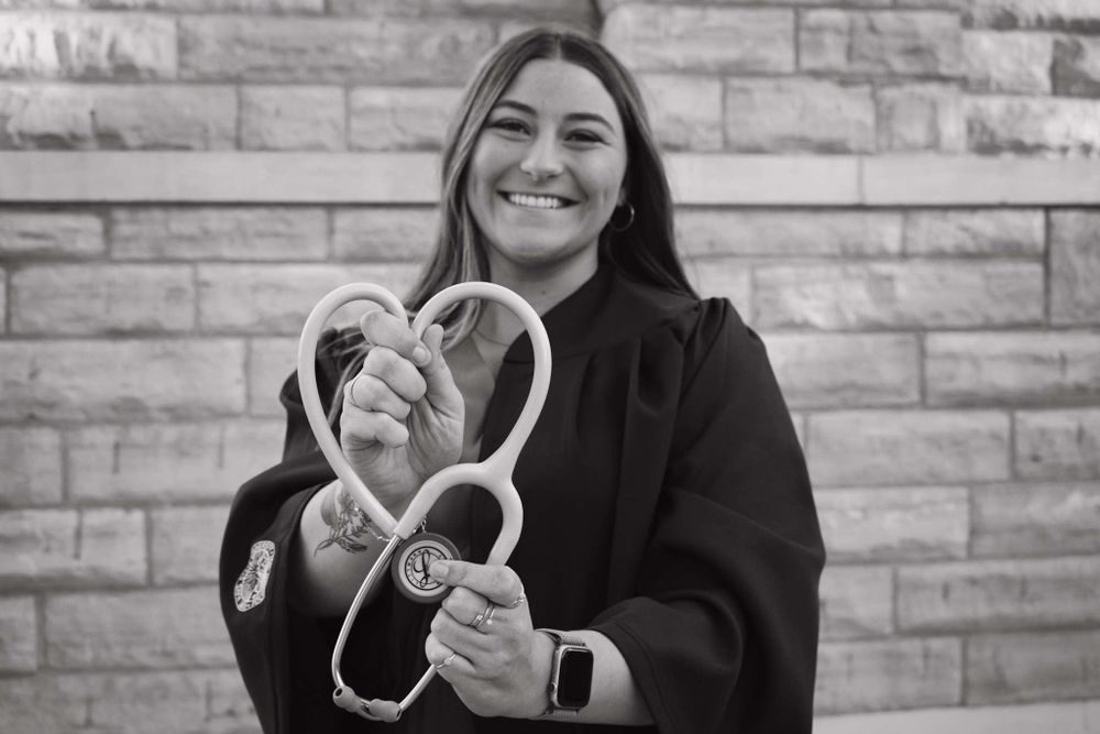 Graduate wearing black gown holds stethoscope shaped into a heart against brick wall.