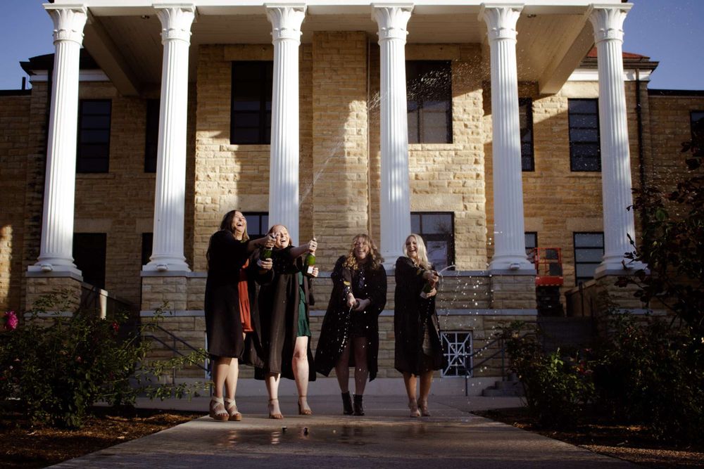 Group of graduates in black gowns pose in front of classical building with white columns.