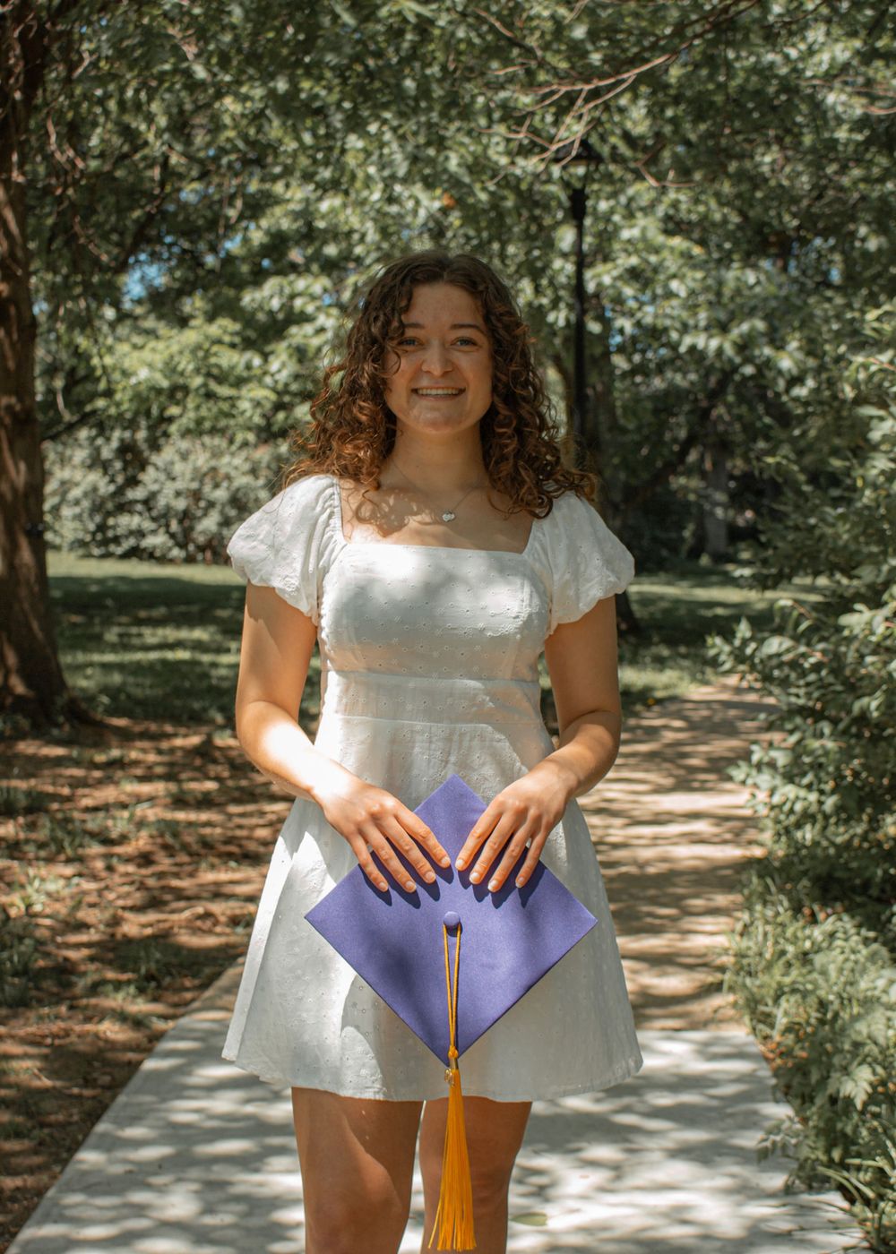 A graduate in a white dress holds purple cap and tassel in a sunlit wooded area.