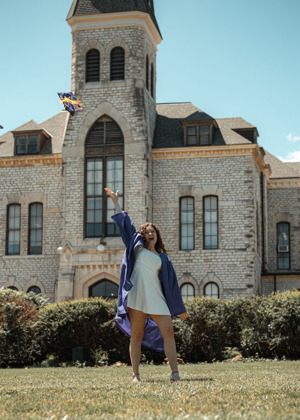 A graduate in blue regalia jumps joyfully in front of a stone university building.