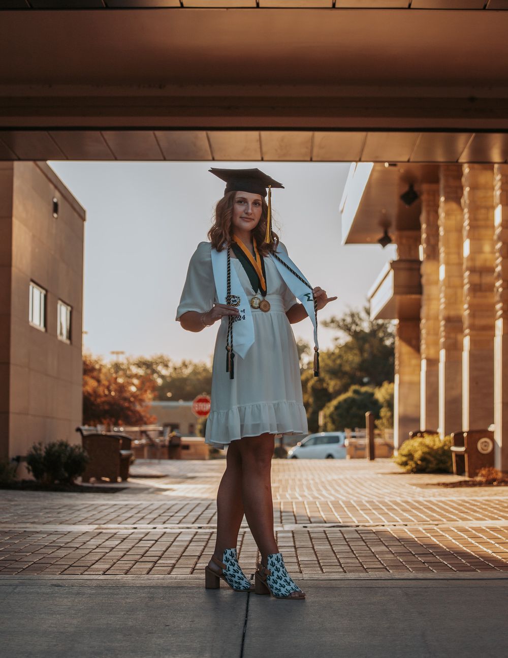 A graduate in light blue regalia poses under a covered walkway at sunset.