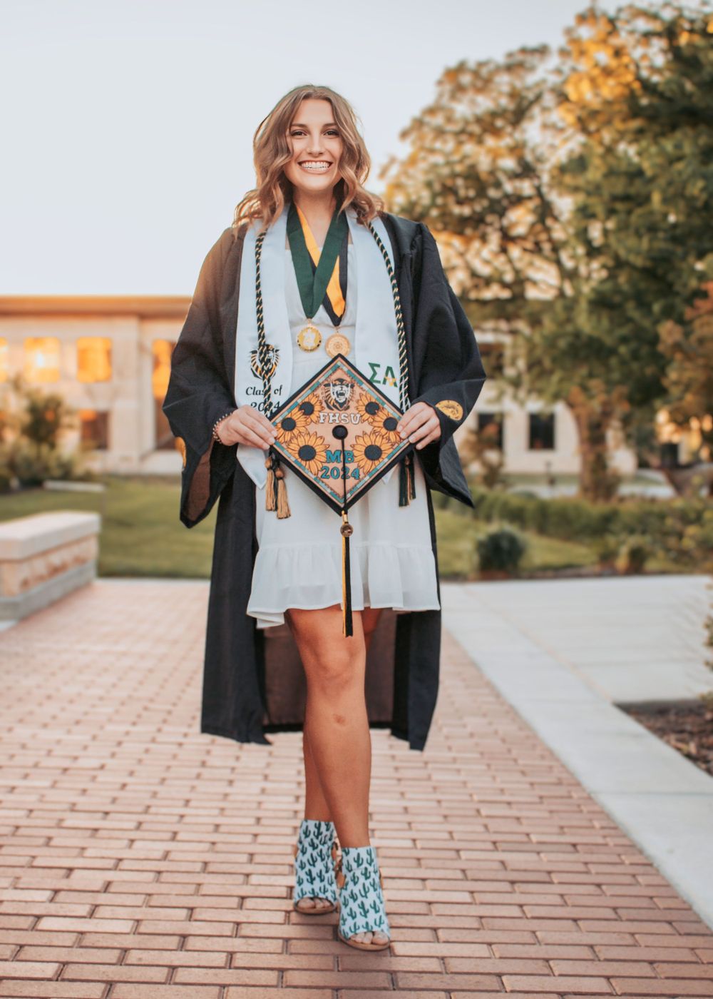 Graduate in black cap and gown poses on brick pathway with decorated graduation cap and patterned boots.