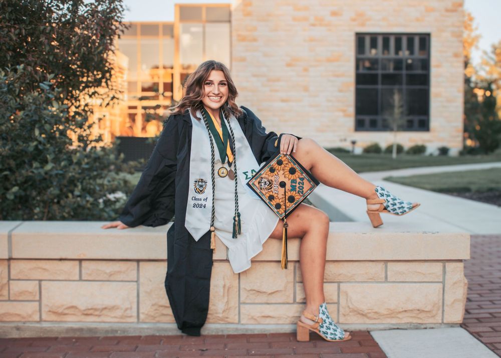 Graduate in black gown sits on stone wall showing decorated cap and patterned shoes.