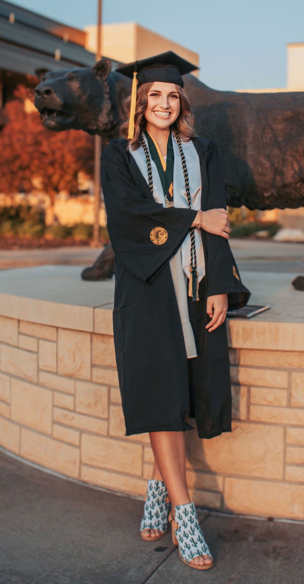 Graduate in black academic regalia poses next to bear statue wearing patterned boots.