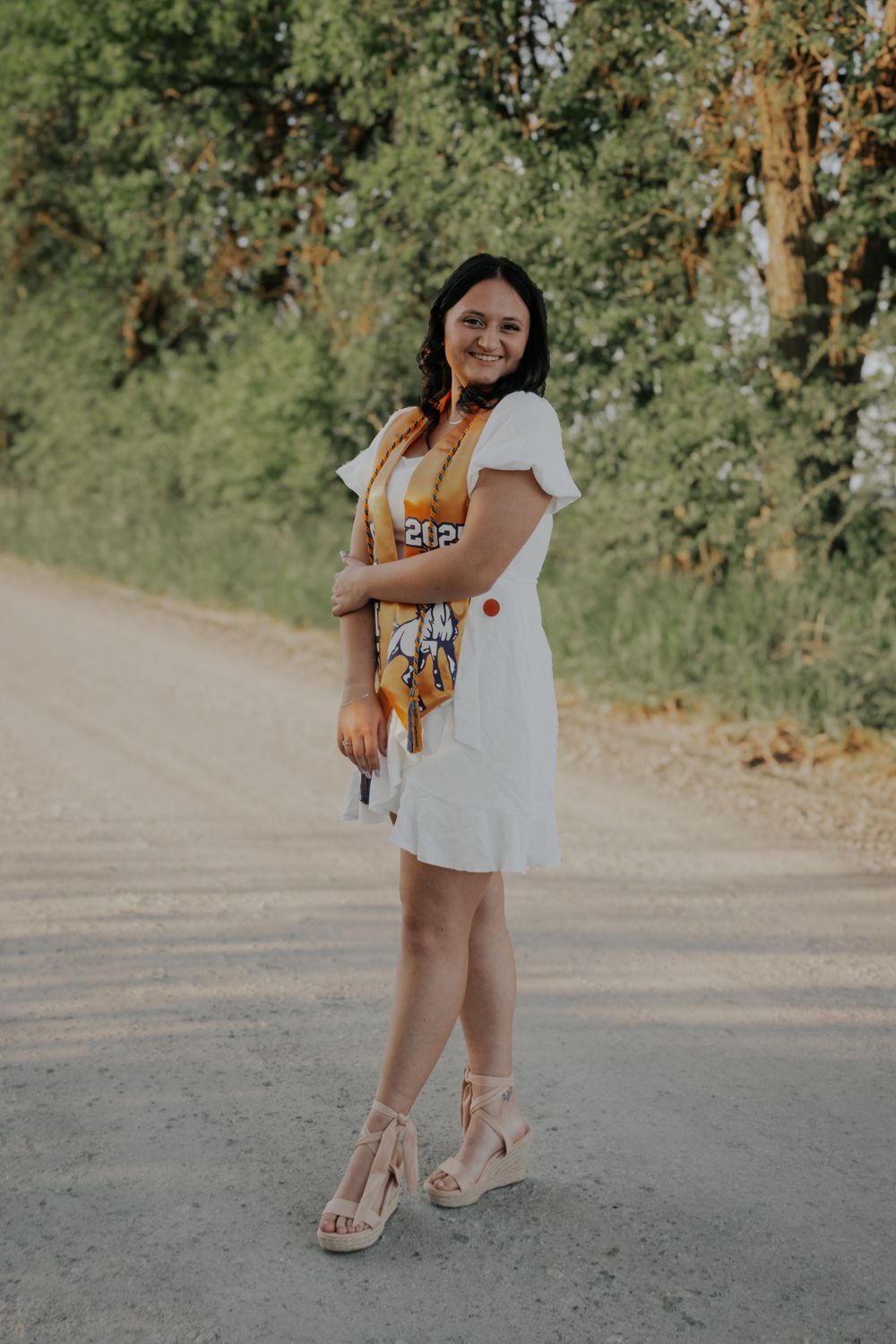 Woman in white summer dress and espadrille wedges poses on dirt path with green trees and foliage in background.