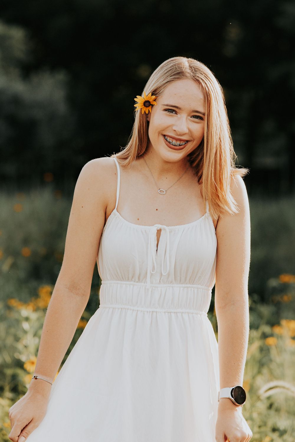A person wearing a white sundress smiles while standing in a field of yellow wildflowers on a sunny day.