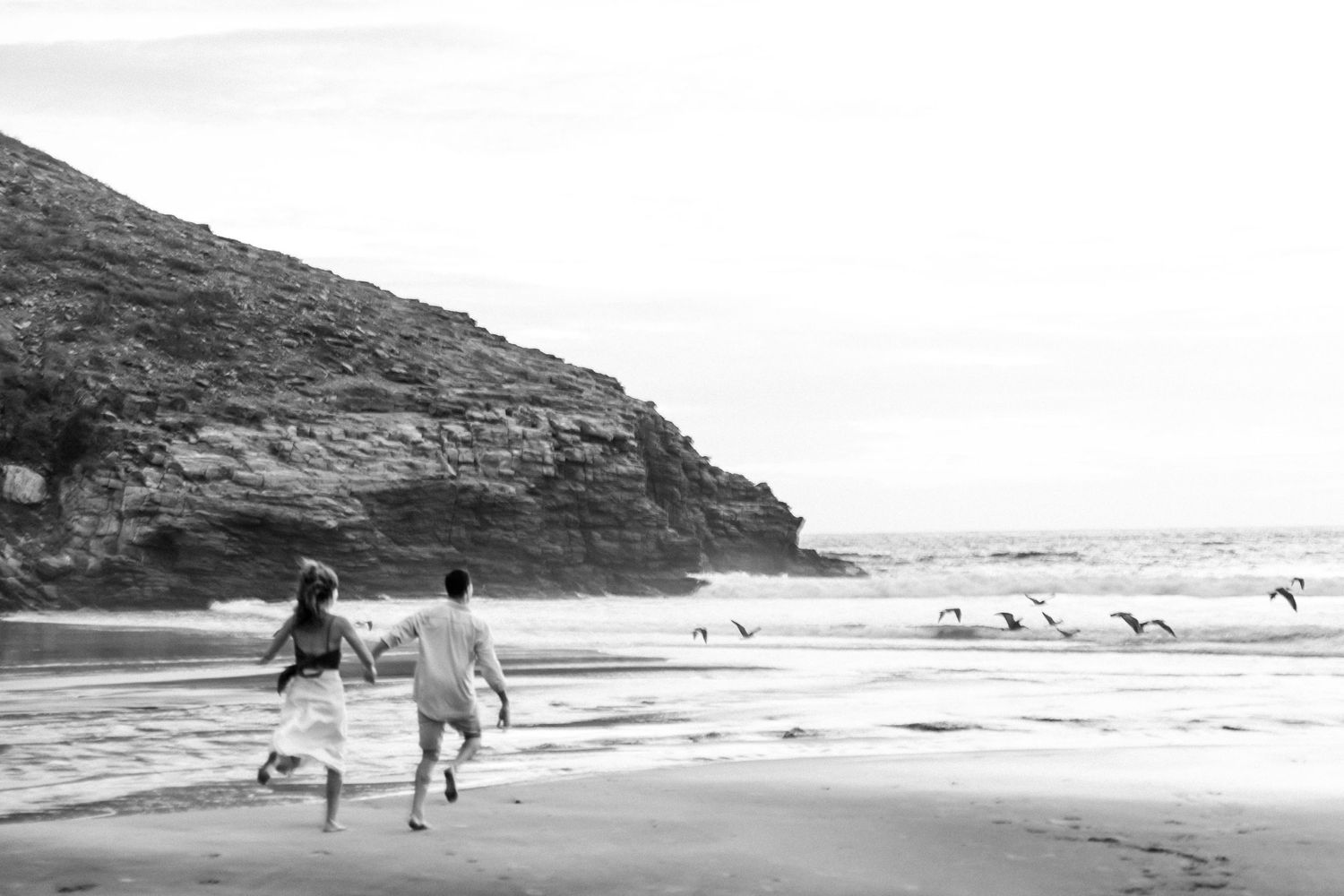 Black and white photo of couple running along beach with seabirds.