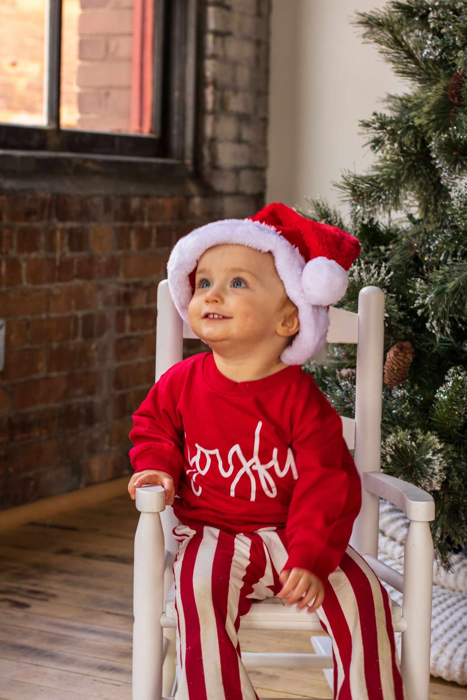 A small child in red and white striped Christmas pajamas and Santa hat sits in a white rocking chair.