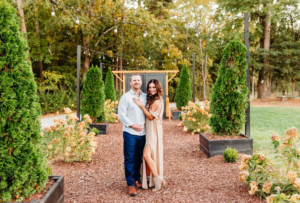 A romantic engagement photoshoot takes place in a lush garden setting with tall trees and mulch-covered paths.