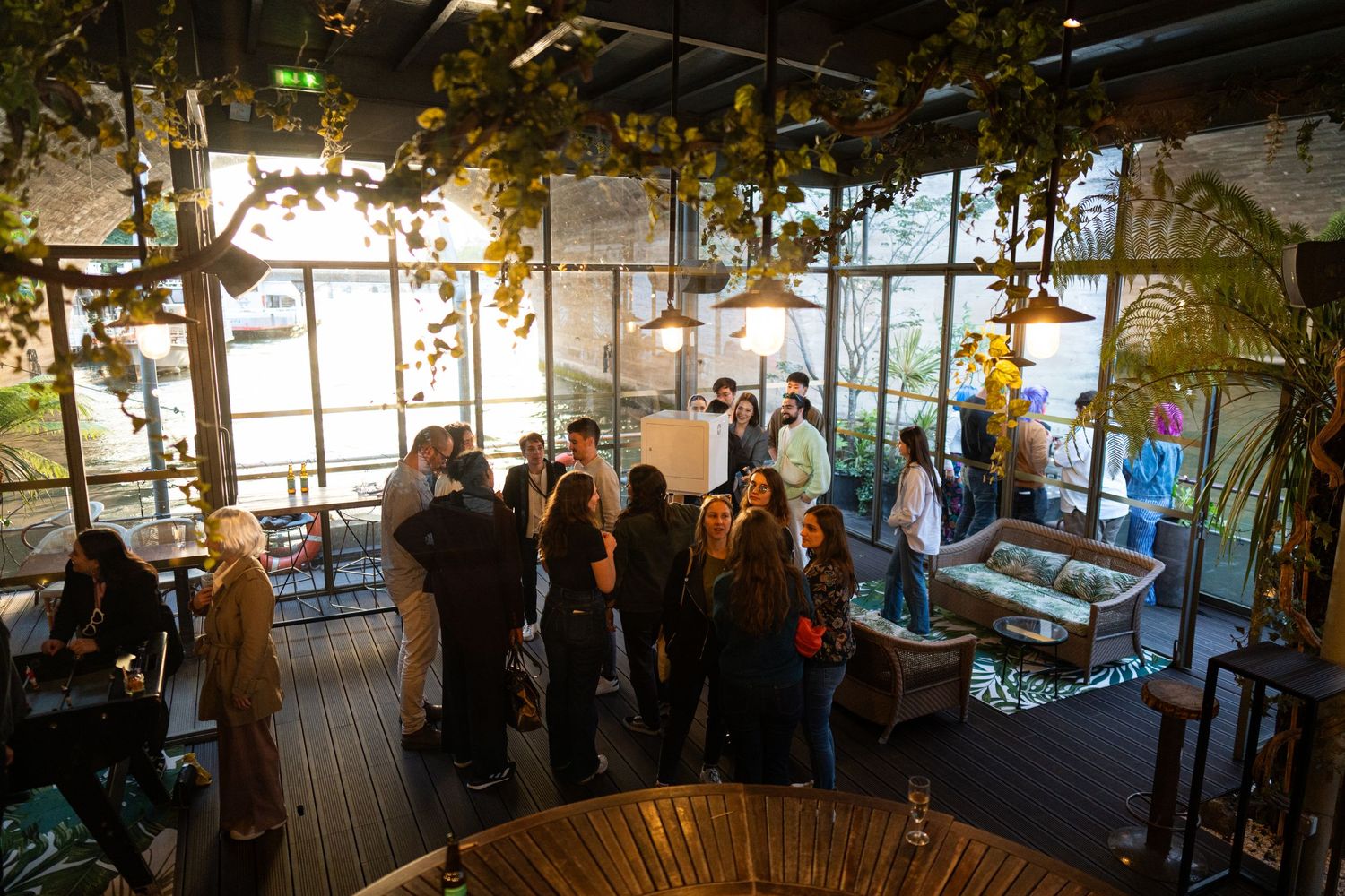 People gather in a sunlit restaurant or cafe space with large windows and hanging plants creating an atmospheric setting.