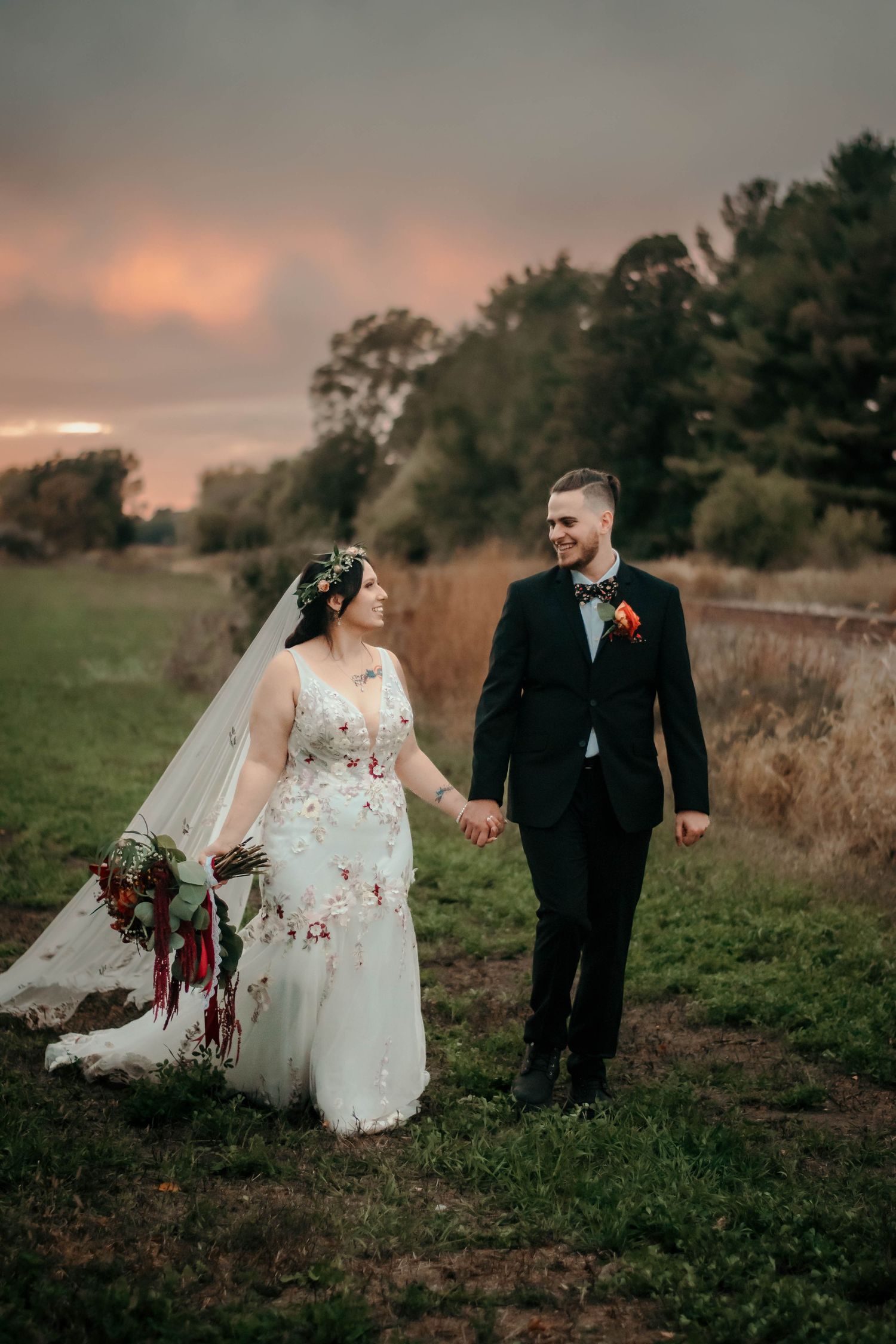 Couple in wedding attire walk together through scenic field during sunset photo session.