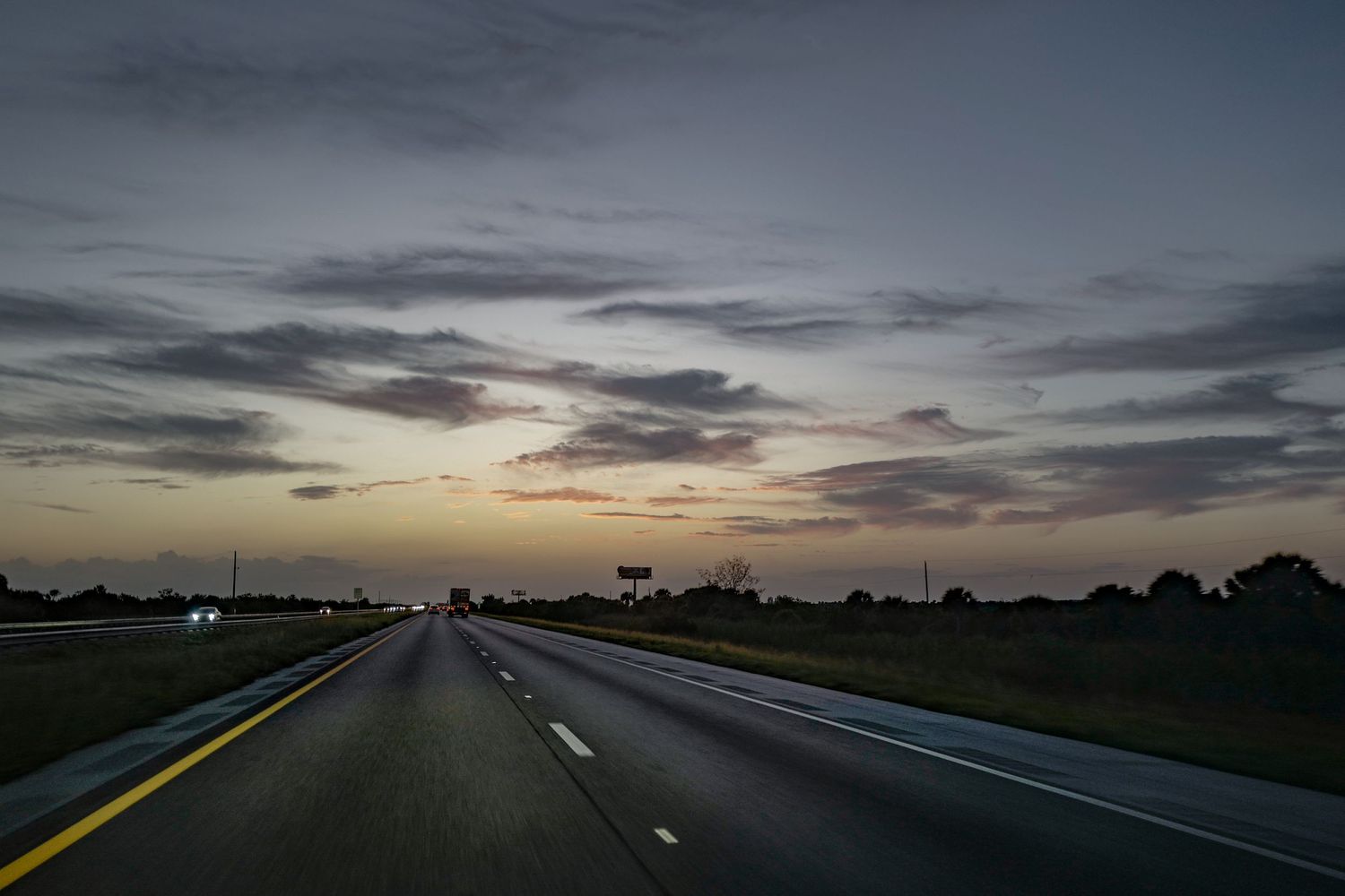 Dark highway stretches into the distance under a dramatic sunset sky with streaks of clouds.