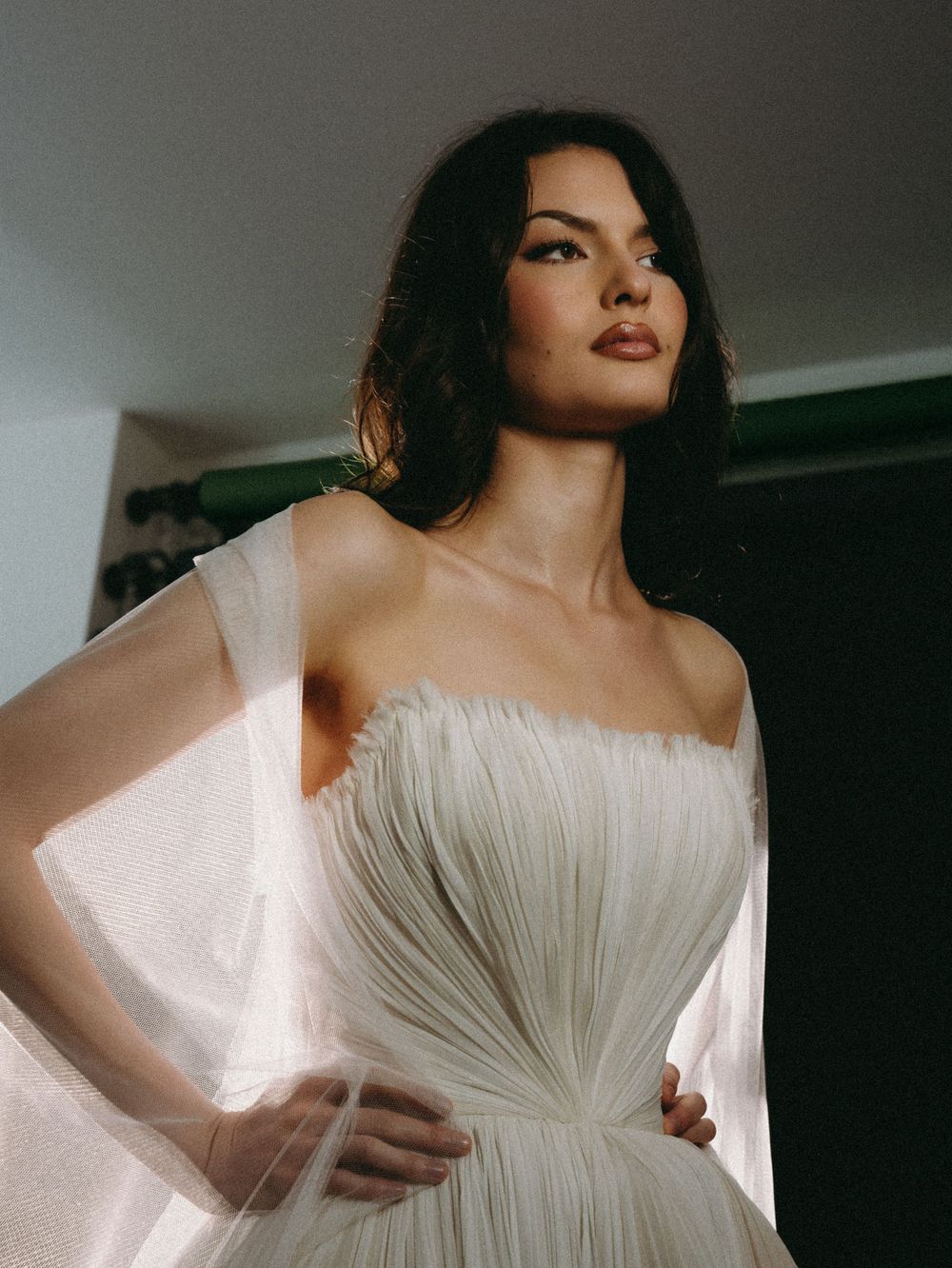 Elegant bride in a strapless pleated white tulle gown with sheer cape sleeves, posing gracefully in soft dramatic lighting.