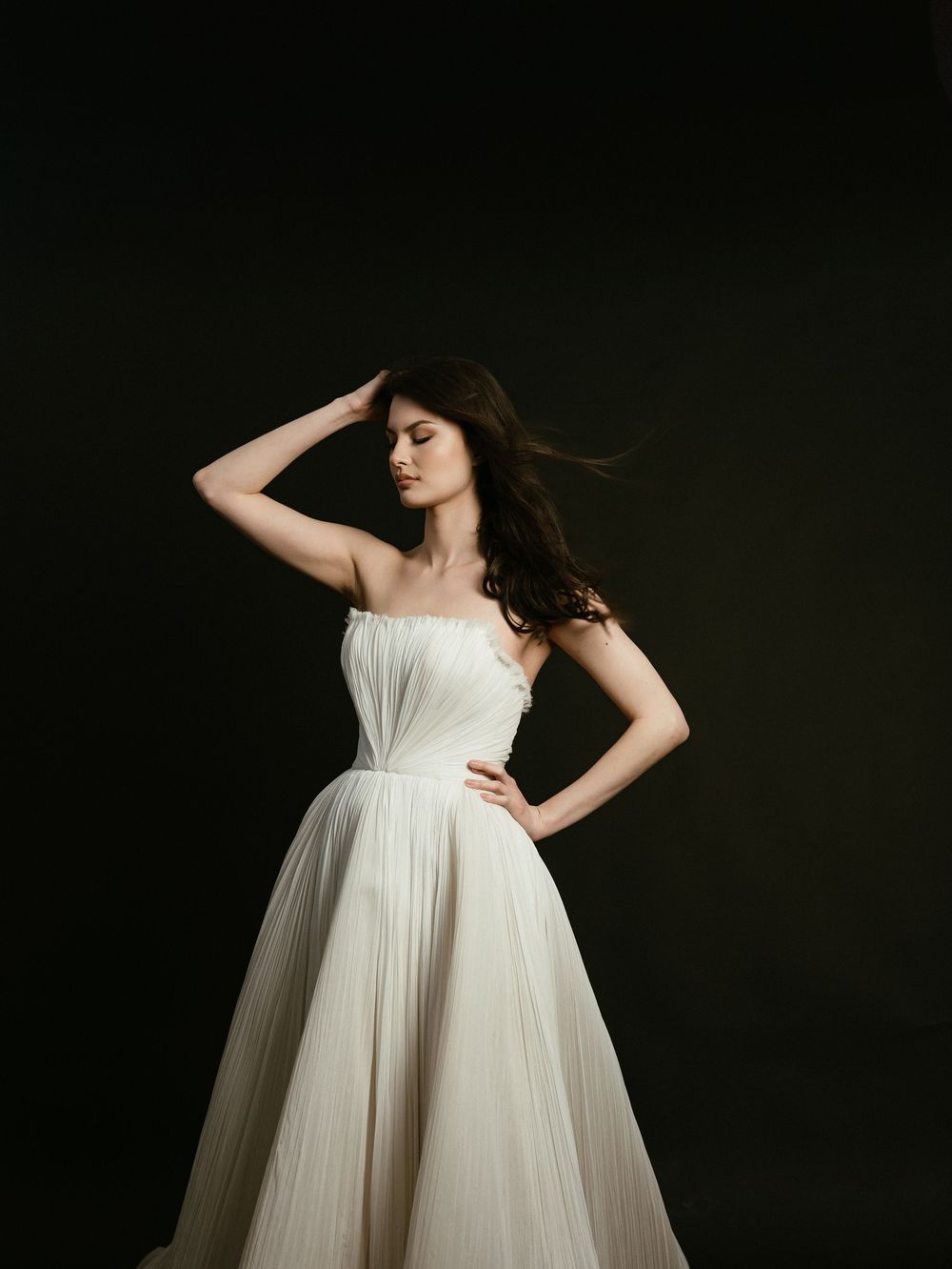 A woman in a strapless white pleated ball gown poses gracefully against a dark background with wind-swept hair.