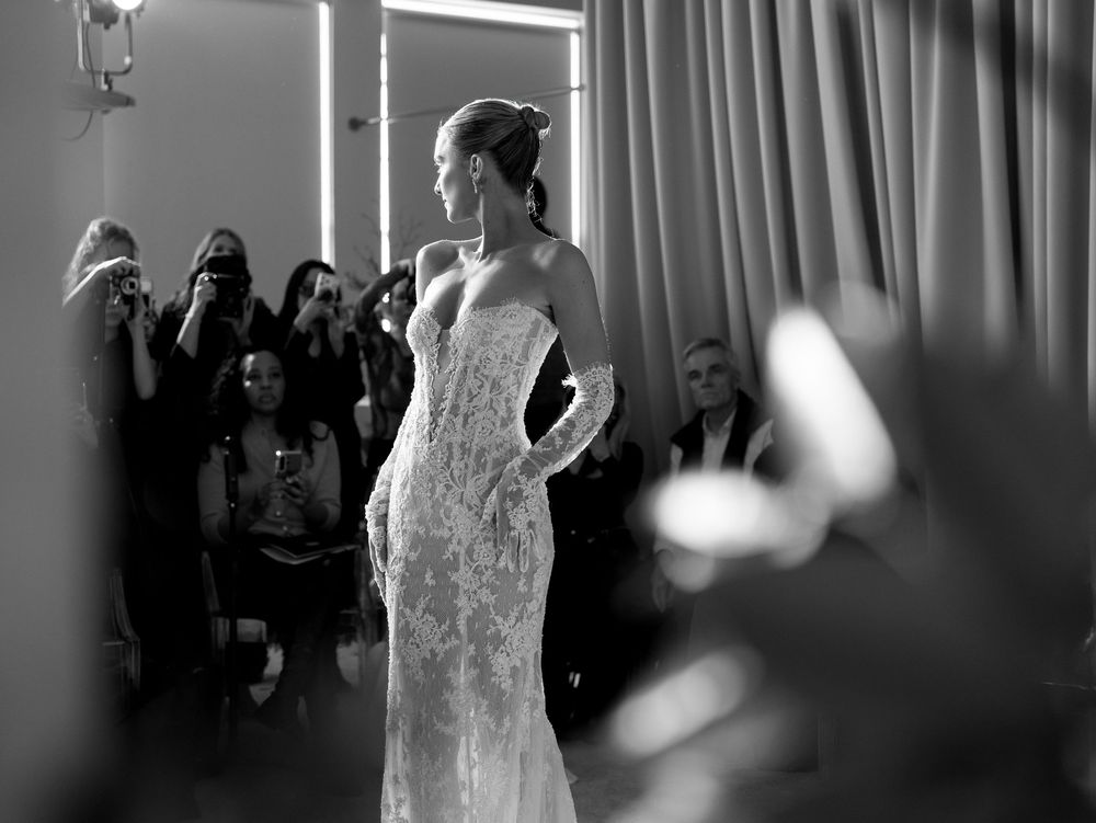 Black and white photo of a bride in a lace gown posing while photographers capture the moment backstage.