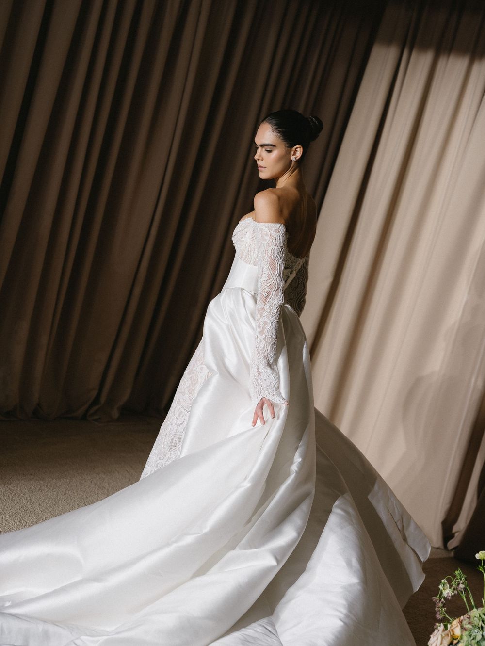 Elegant bride in an off-shoulder lace and satin ballgown with a long train, posing gracefully in soft lighting.
