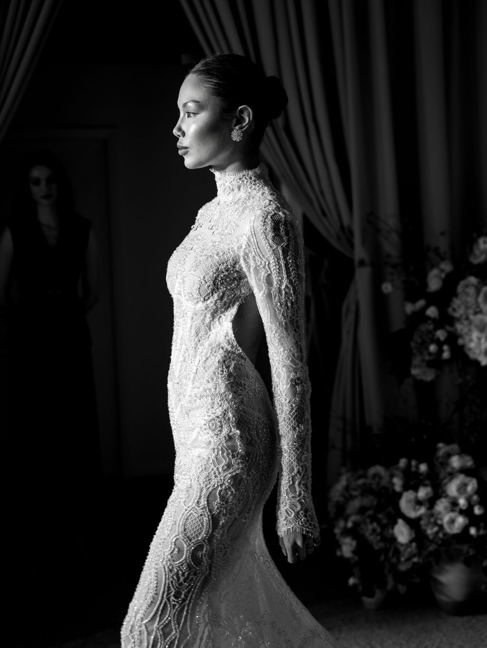 Black and white photo of a model in an elegant lace high-neck gown with long sleeves, posed in profile.