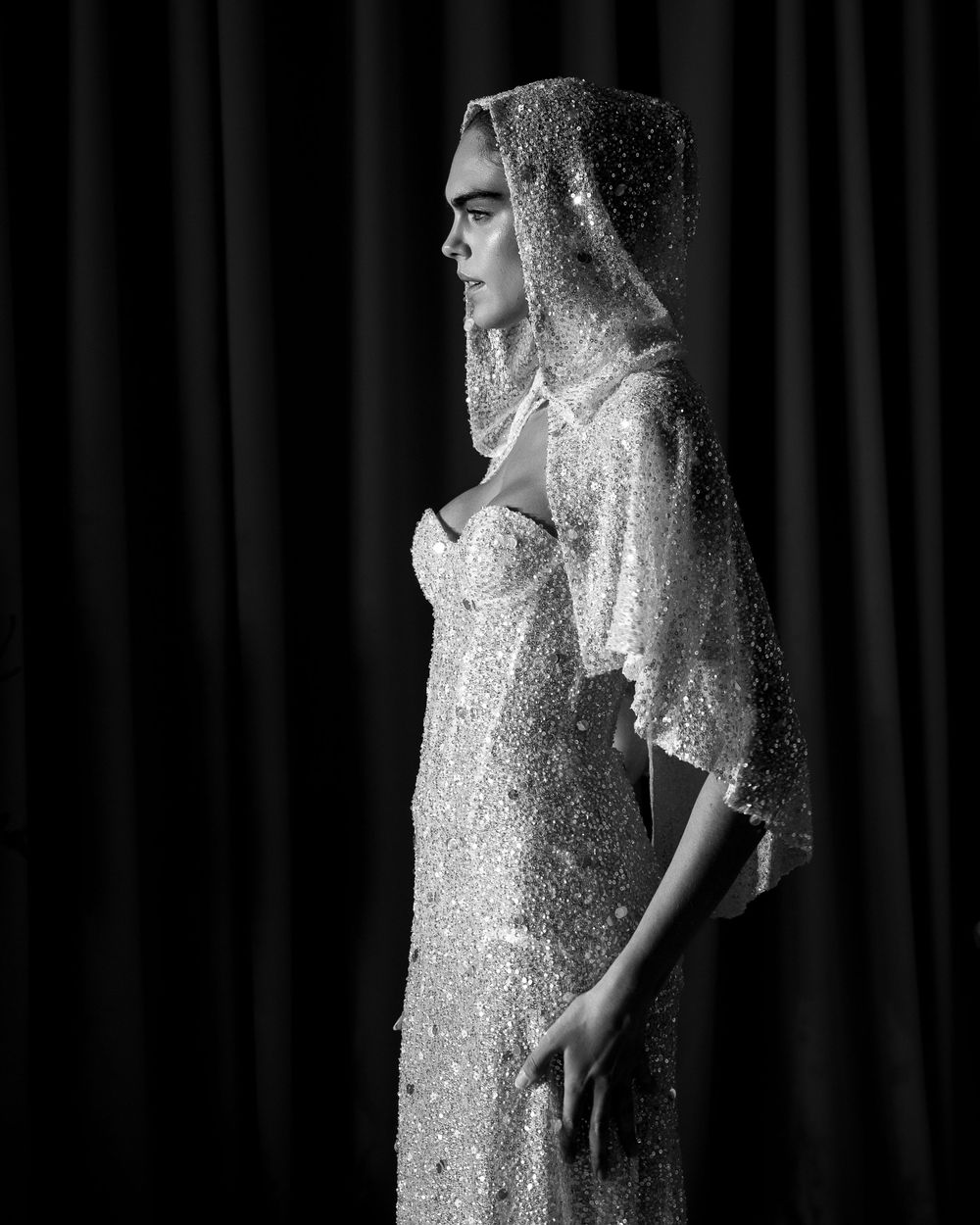 Black and white photo of a model in a sequined gown and veil, posing elegantly against dark curtains.