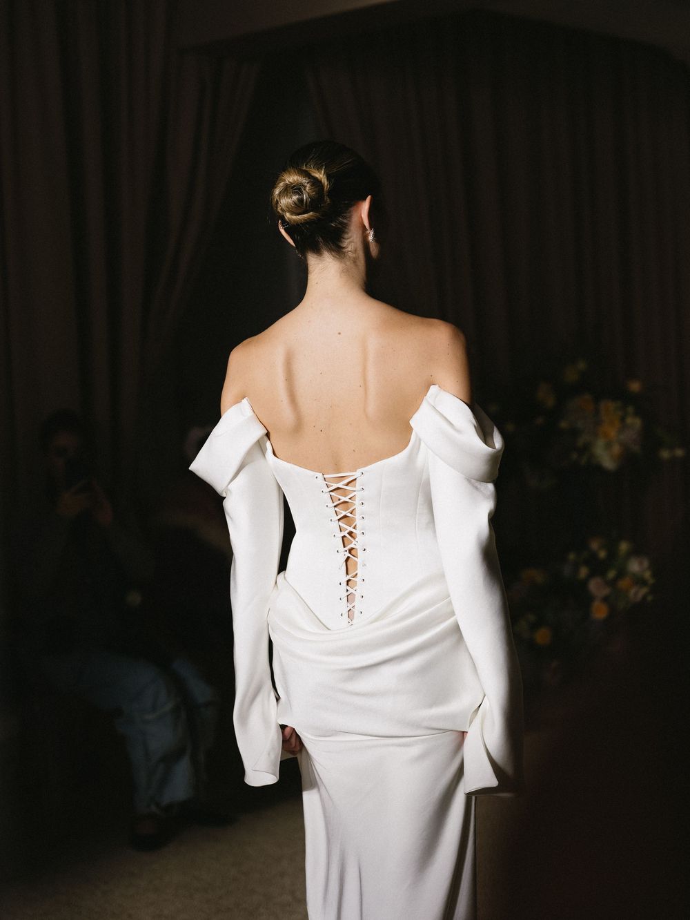 Back view of a bride in a white off-shoulder gown with lace-up corset detail and long sleeves.