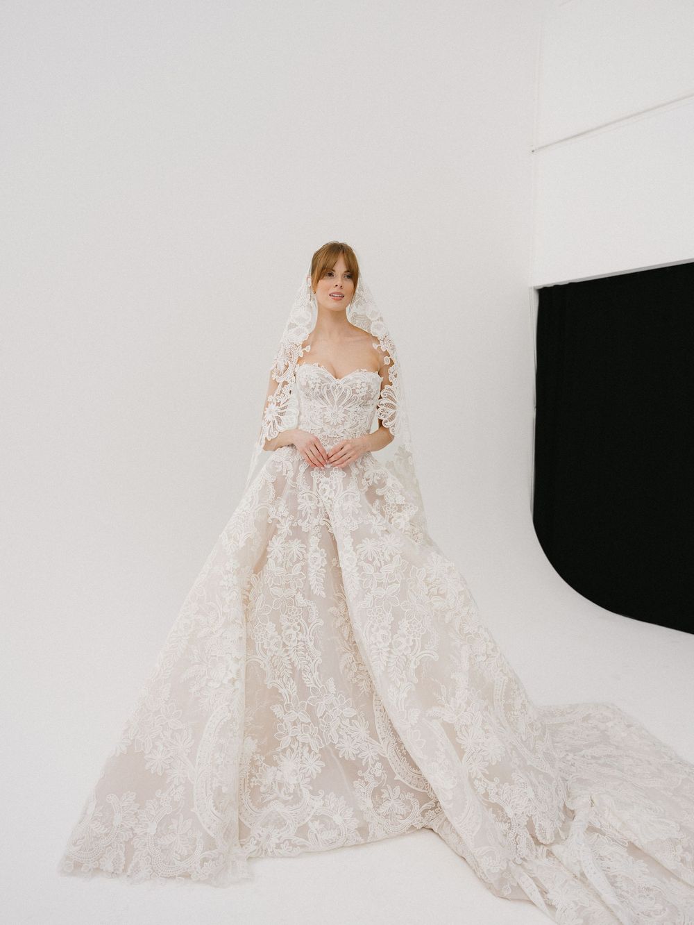 Model wearing an elaborate ivory lace ball gown wedding dress with long sleeves and an extended train in a studio.