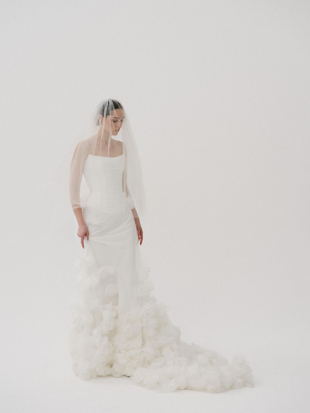 Elegant bride in a white fitted gown with ruffled train and sheer veil, posing gracefully against a white background.