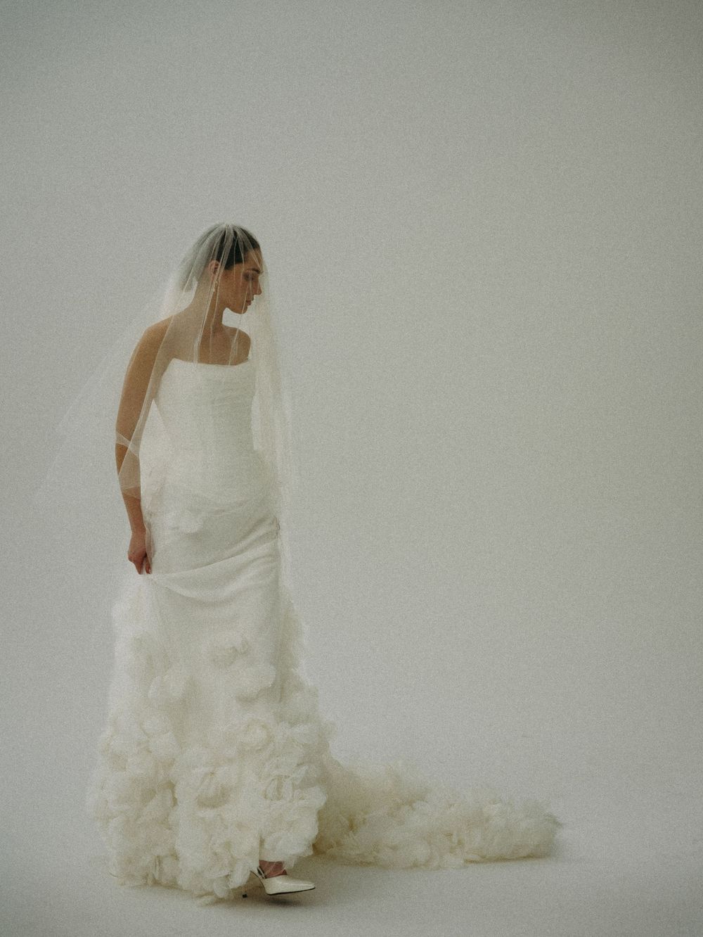 Bride in a strapless white ruffled gown with a flowing veil, posing elegantly against a soft white background.