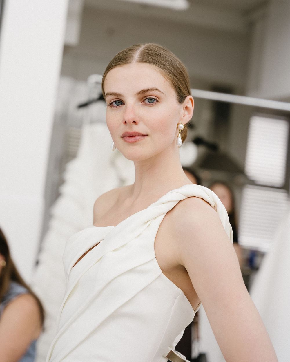 A model in a white one-shoulder draped gown with gold earrings poses elegantly in a bright, minimalist studio setting.