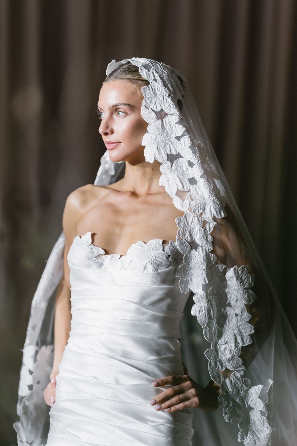 Elegant bride in strapless white gown with floral lace veil, posing gracefully in soft studio lighting.