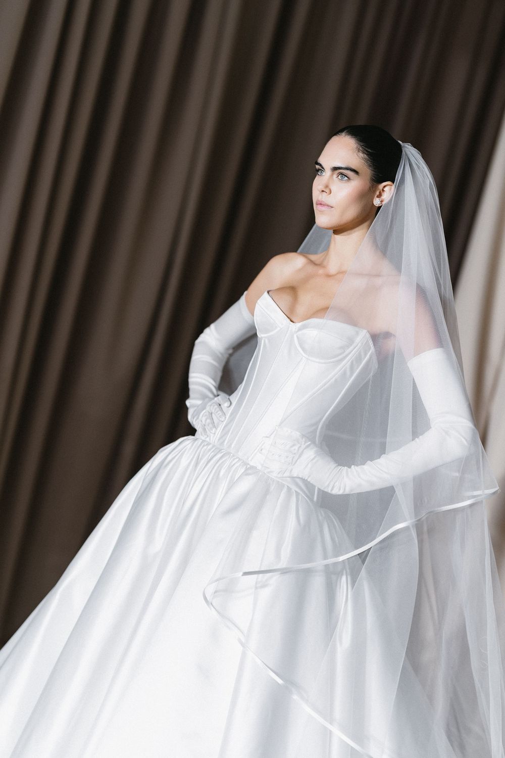 Elegant bride in white strapless corset gown with full skirt, long gloves, and veil, posing gracefully indoors.