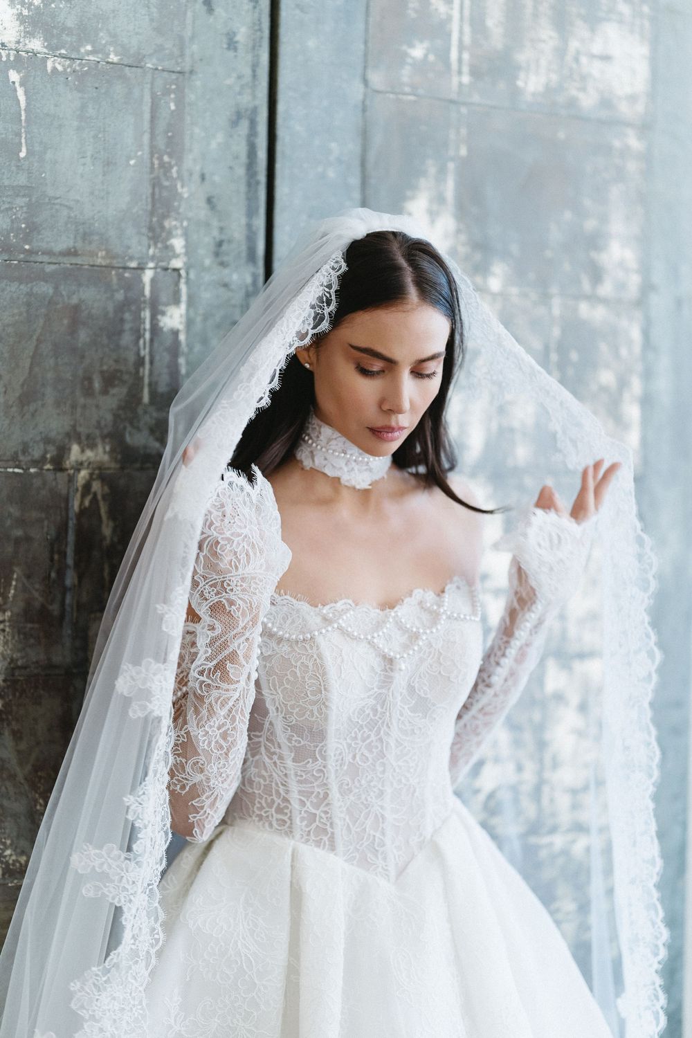 Elegant bride in lace wedding gown and veil poses gracefully against a rustic gray wall.