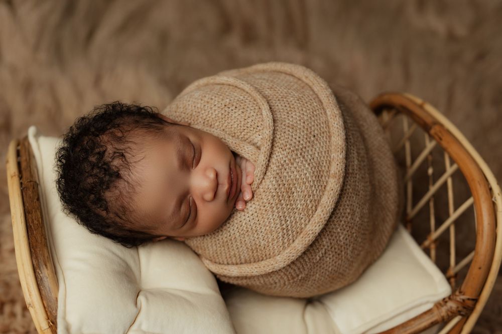 Newborn baby sleeping peacefully in beige knit wrap on white pillow in wooden basket.