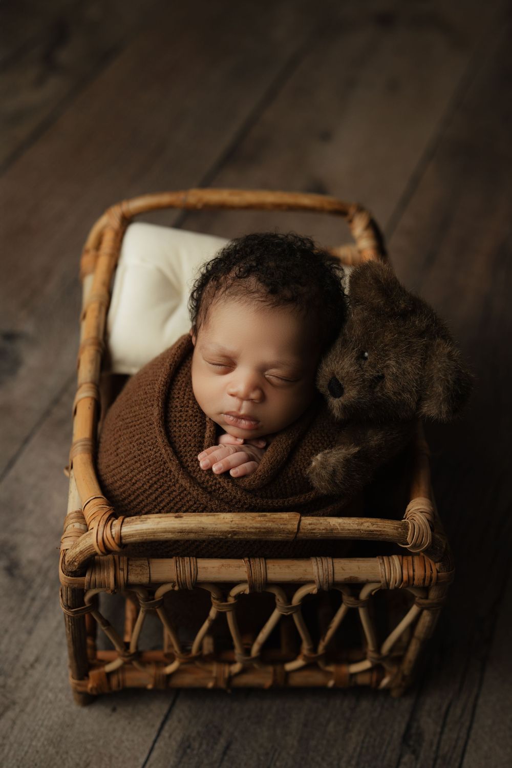 A sleeping newborn in brown knit wrap rests peacefully in a vintage woven wicker bed on dark wooden floor.