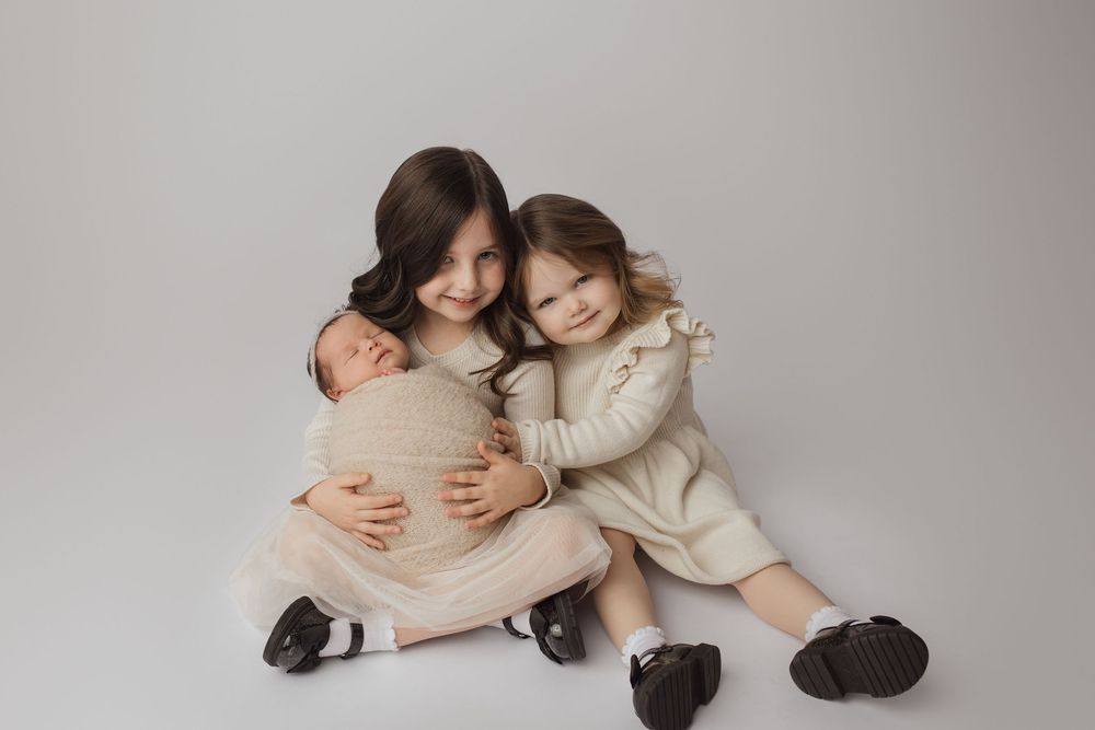 Two children in light colored dresses sit together on a light gray backdrop wearing matching brown shoes.