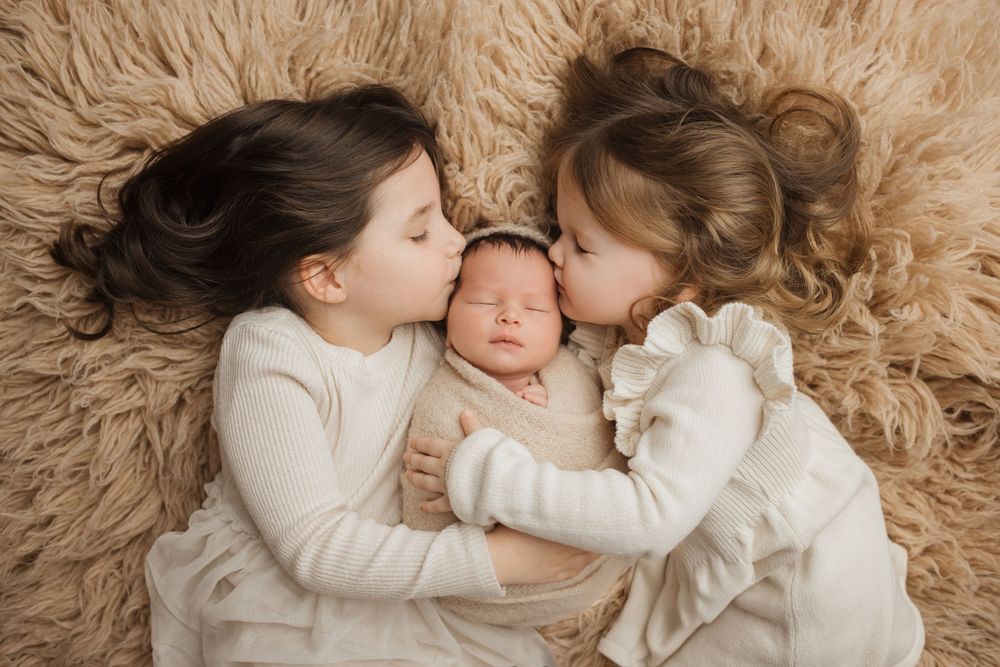 Two children snuggling with a sleeping newborn baby on soft beige fur backdrop in tender family moment.