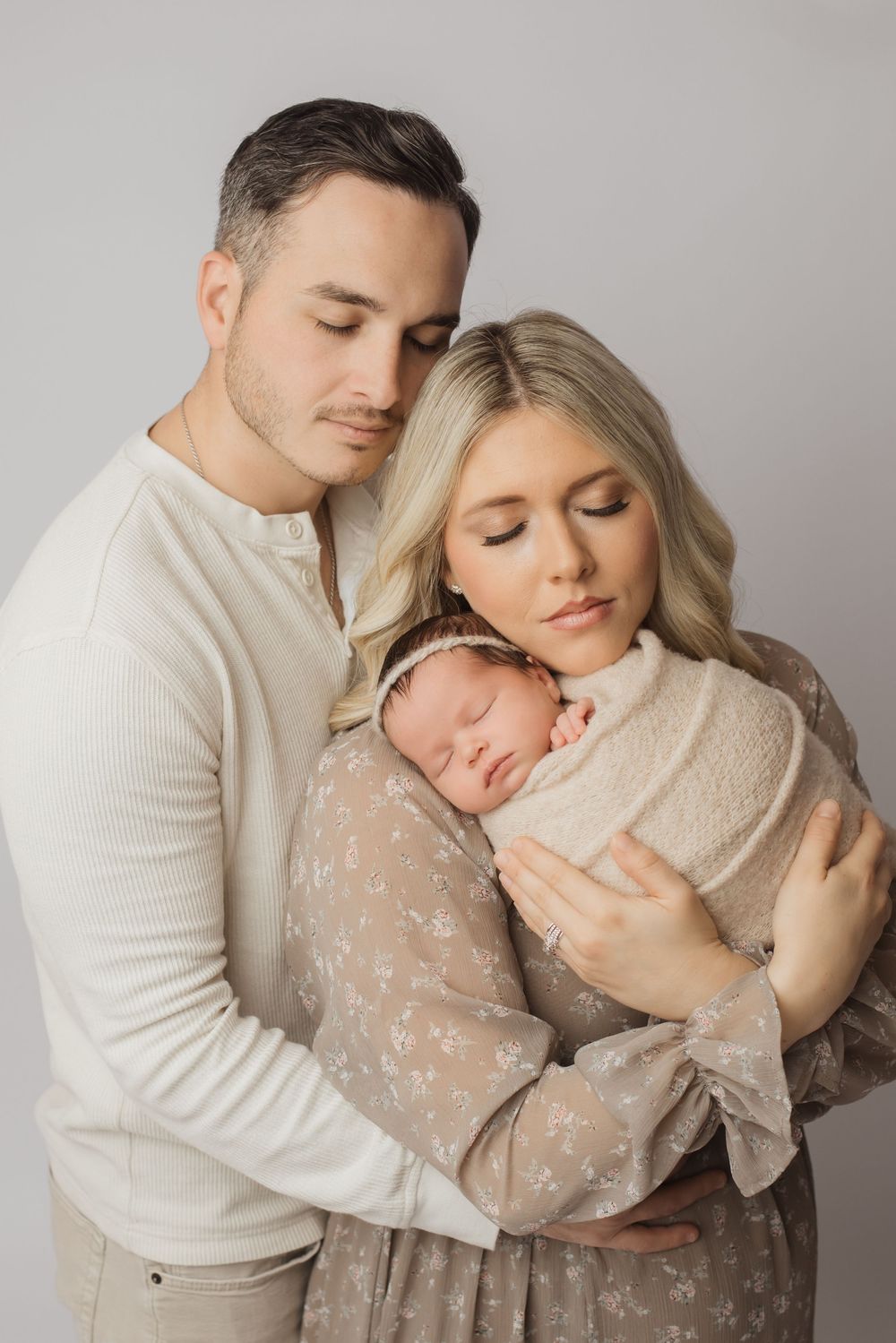 A couple in cream colored outfits tenderly holds their sleeping newborn baby against a neutral background.