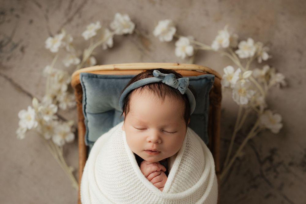 A sleeping baby wrapped in white swaddling cloth with delicate white floral decorations in the background.