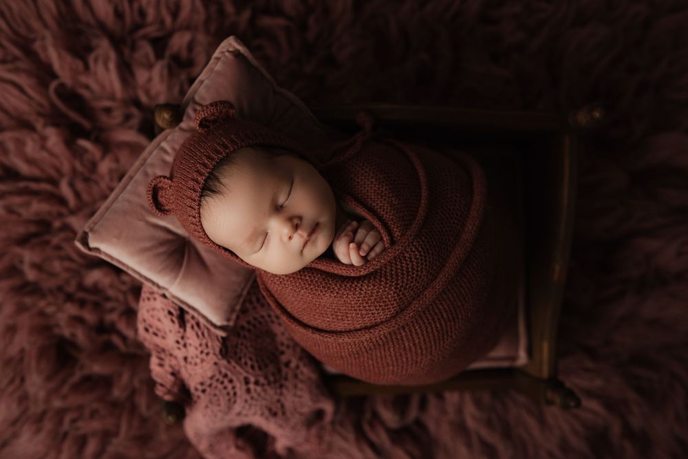 Sleeping newborn wrapped in brown knit swaddle resting on textured burgundy blanket in moody lighting.