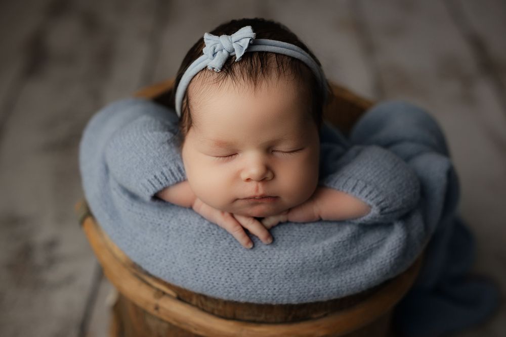 A sleeping newborn baby rests peacefully on a gray pillow in a rustic wooden basket.