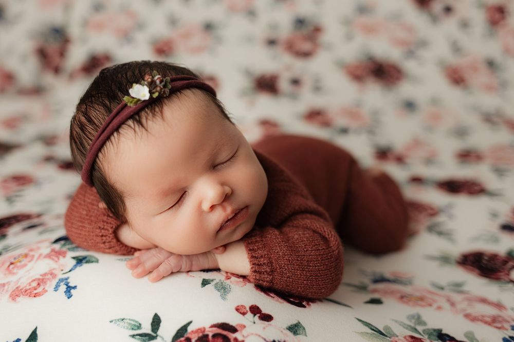 Sleeping newborn baby wrapped in burgundy swaddle lying on floral pattern blanket with soft natural lighting.