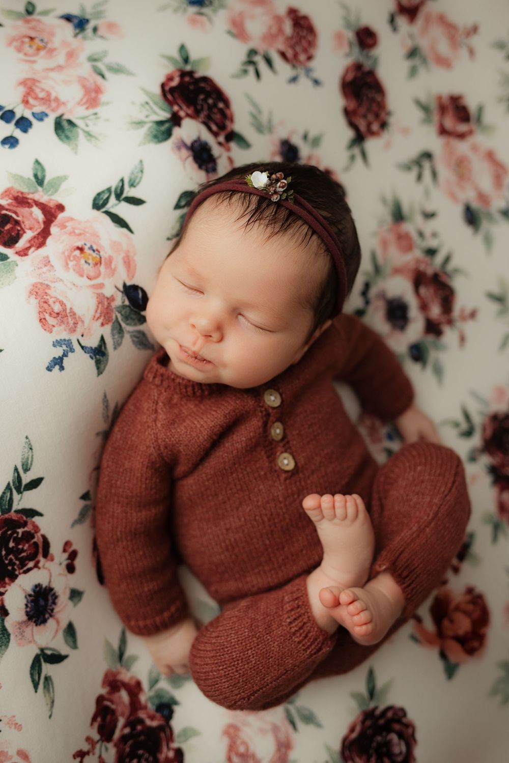 Sleeping newborn in rusty brown knit outfit lies on floral pattern backdrop wearing delicate headband.
