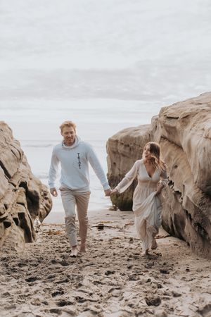 Couple runs playfully between rock formations on cloudy beach day.