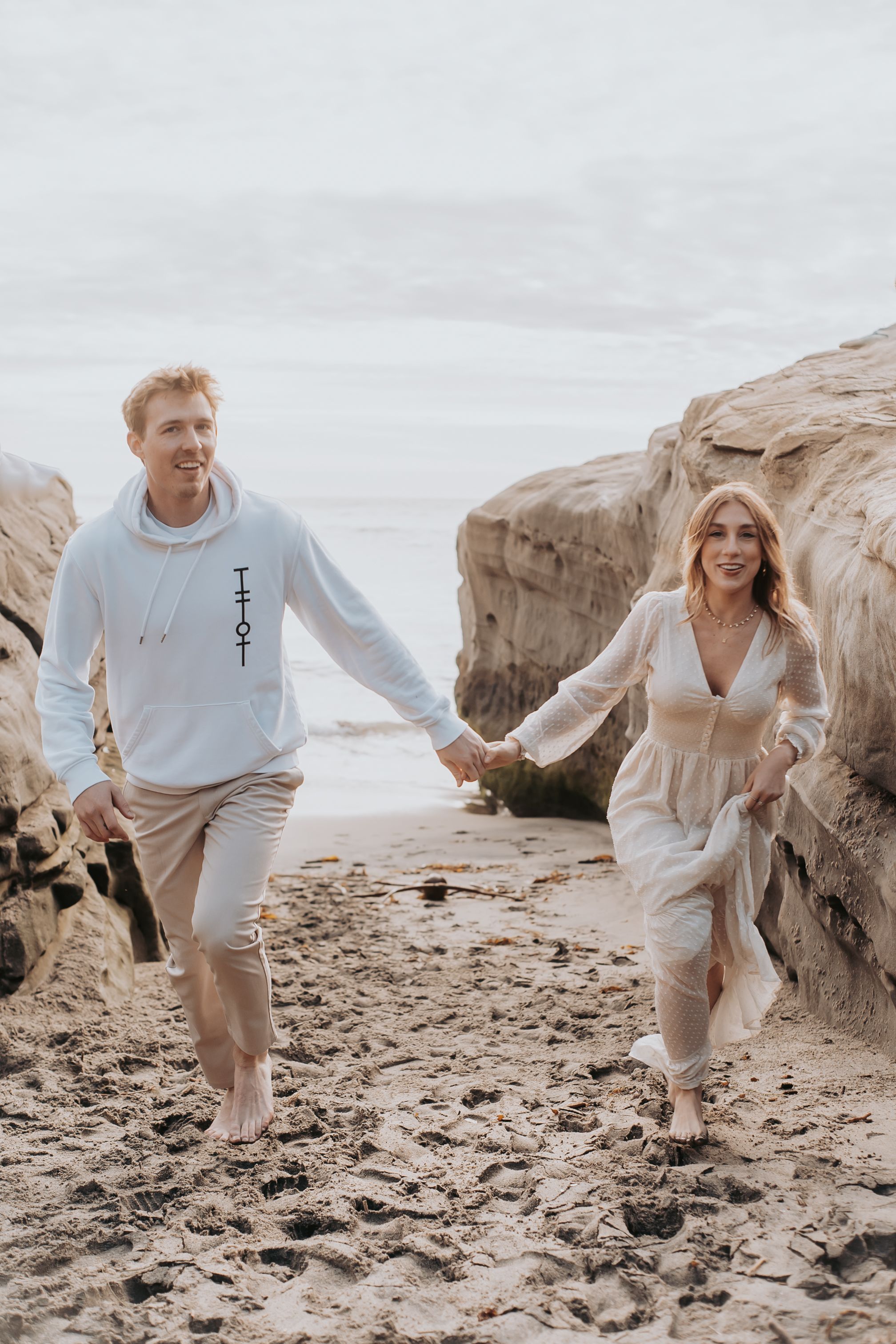 Couple in light colored outfits playfully runs along rocky beach coastline.