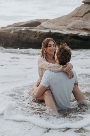 A couple embraces playfully in the surf near rocky coastal cliffs.