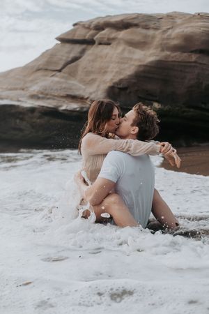 A couple shares an intimate moment while being splashed by ocean waves near rocky cliffs.