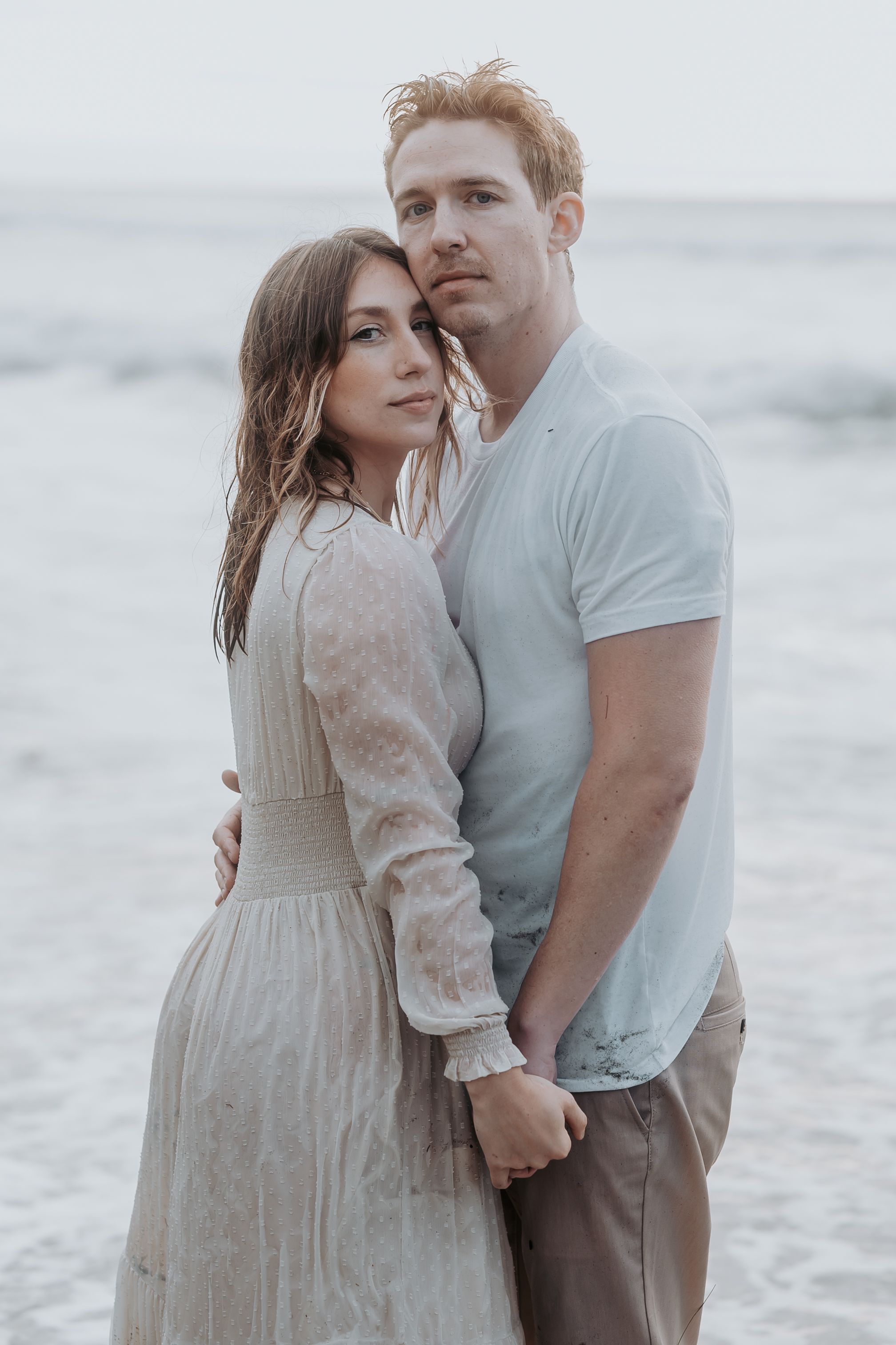 A couple in vintage-inspired clothing embraces by the ocean in soft lighting.